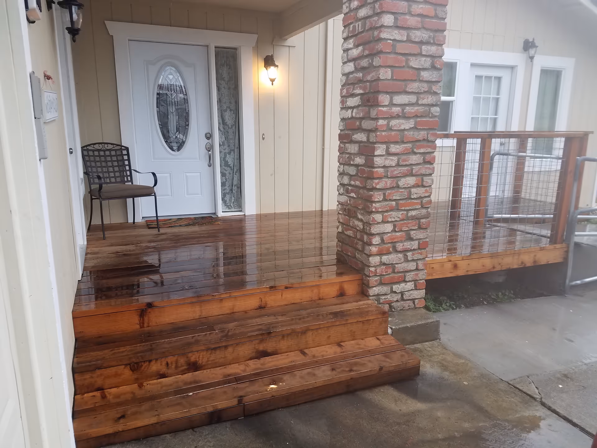 Front porch area of a building with wet wooden steps and deck, a white door with an oval glass window, a metal chair with a cushion, a brick pillar, and a railing with wire mesh. The porch light is on and the ground is wet from rain.