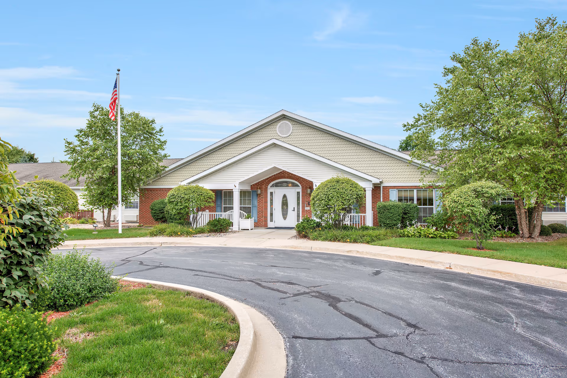 Front exterior of a single-story senior living facility with a circular driveway, landscaped shrubs, and an American flagpole.