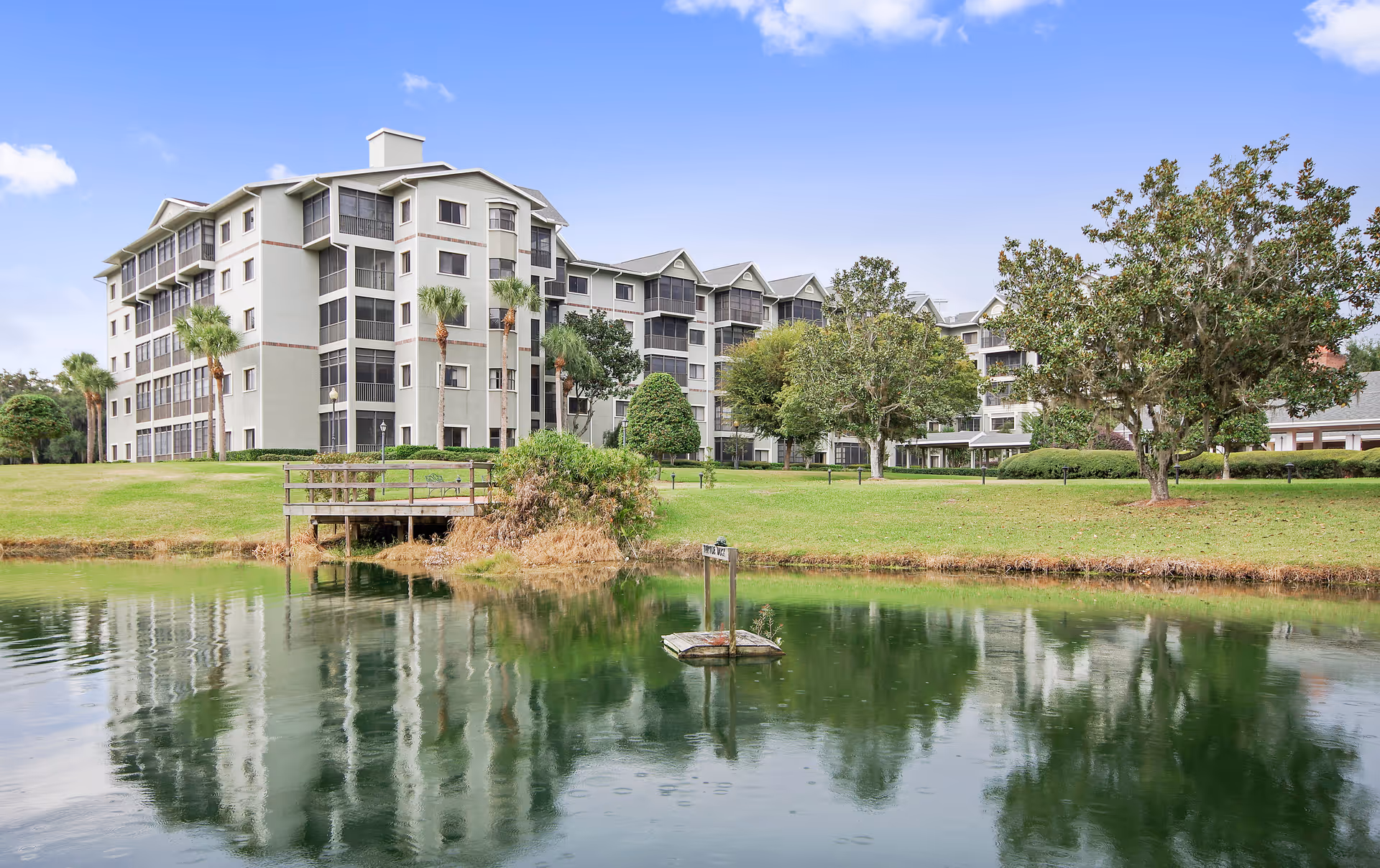Large multi-story senior living building sits beside a pond with trees and green lawn.