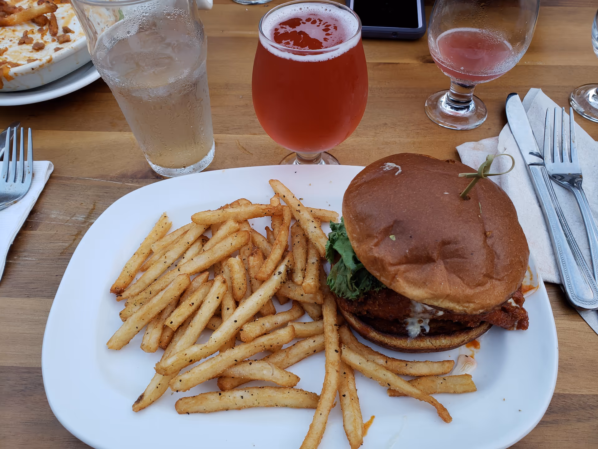 Burger with fries on a white plate, surrounded by drinks on a wooden dining table.