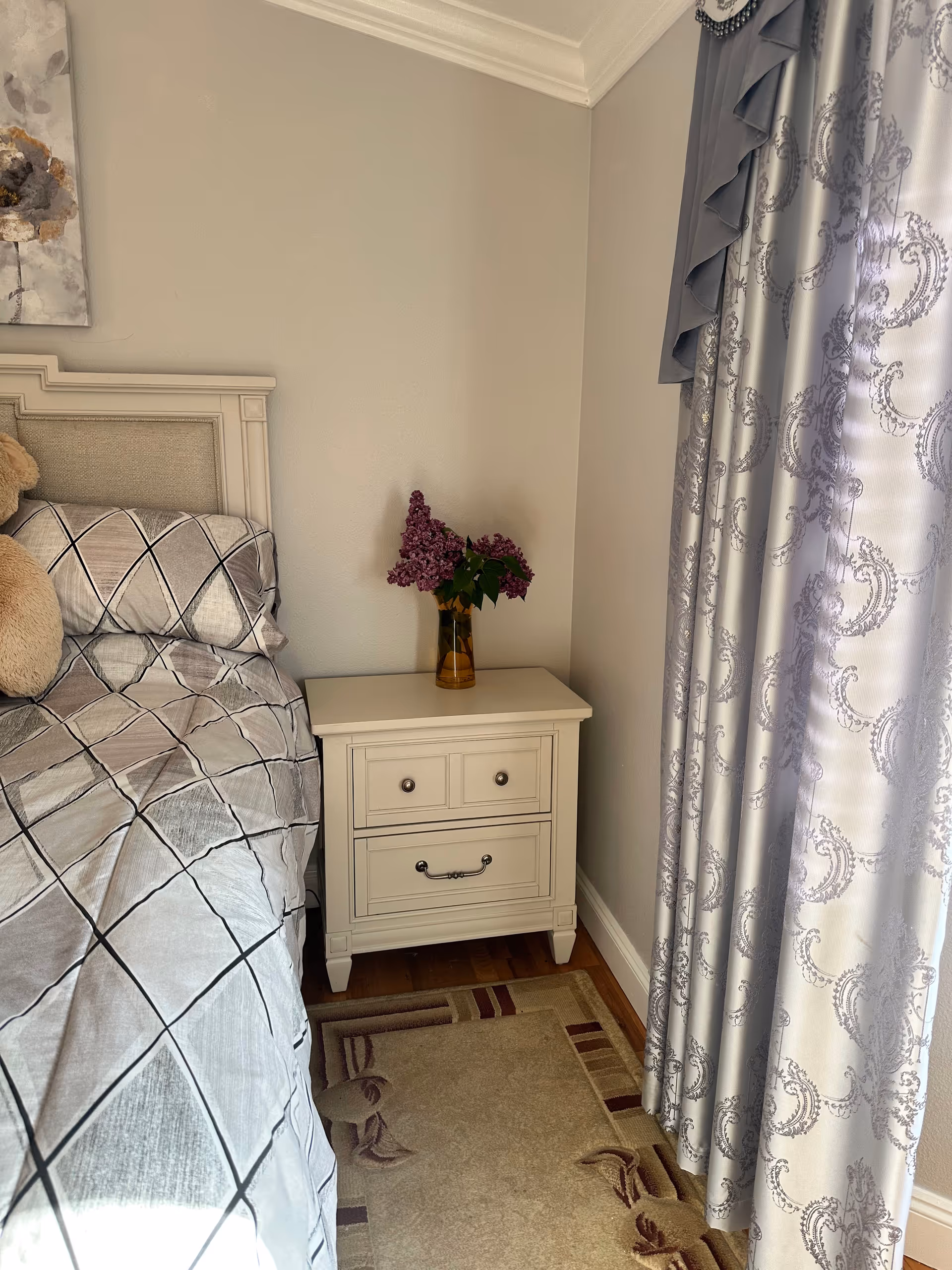 Cozy bedroom with a bed, nightstand, and vase of purple flowers.