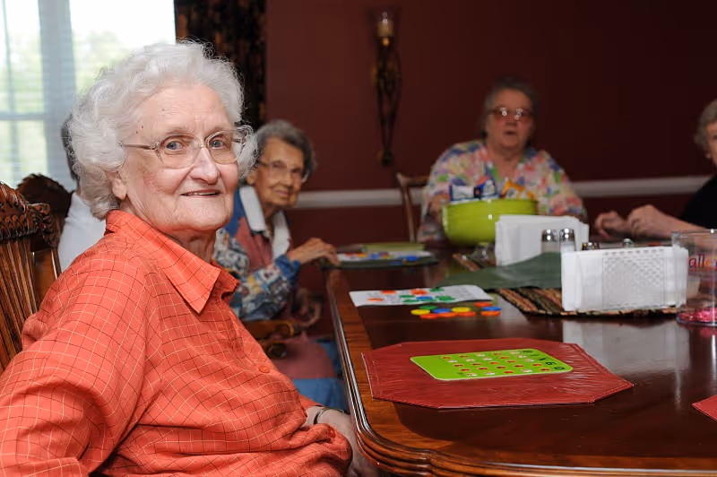 A group of elderly women sitting around a wooden table playing a board game in a warmly lit room with red walls and a window with blinds in the background.