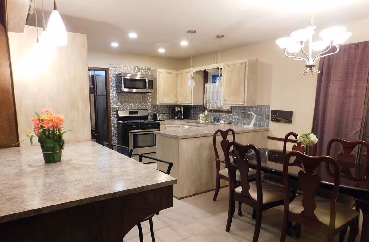 Interior view of a kitchen and dining area with beige cabinets, stainless steel appliances including a microwave and stove, a kitchen island, and a dining table with six wooden chairs. The room is well-lit with ceiling lights and pendant lights, and there are flowers on the kitchen counter and dining table.