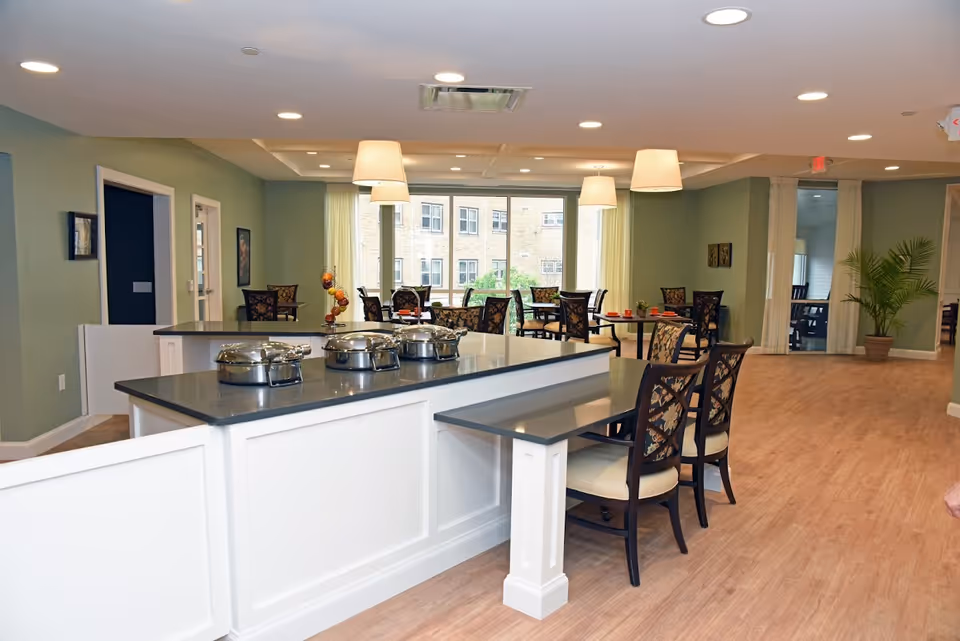 Open dining area with a white kitchen island, upholstered chairs, and tables by large windows.