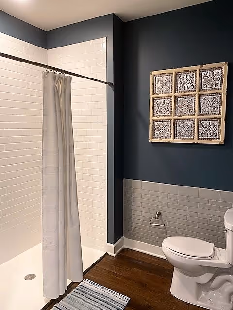 Bathroom with a white tiled shower area and a white shower curtain on a rod. The walls are painted dark blue with a section of gray subway tiles. There is a white toilet and a decorative wall art piece with nine square tiles above the toilet. A striped rug is on the wooden floor in front of the shower.