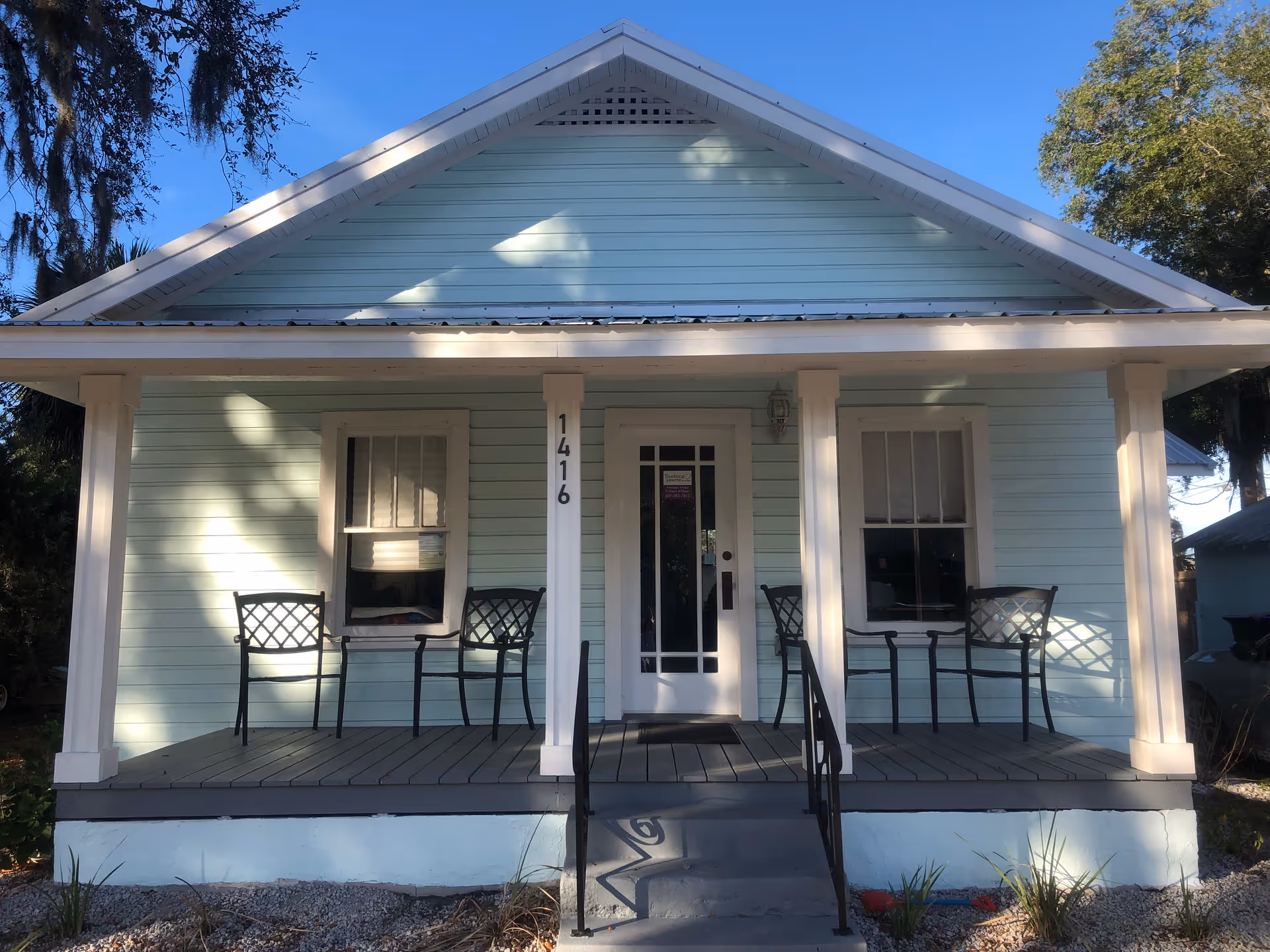 Light blue house with white trim and a front porch, four chairs, and steps.