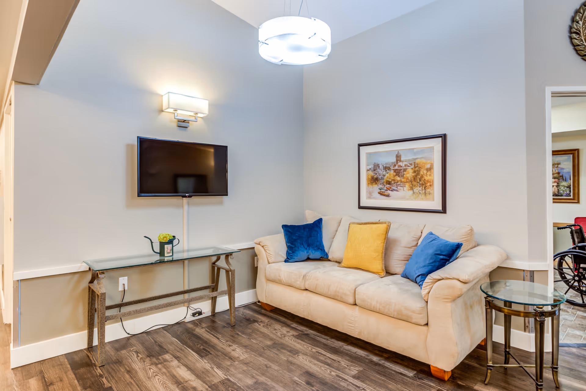 Cozy living room in assisted living facility with beige sofa, wall art, TV, and glass tables.