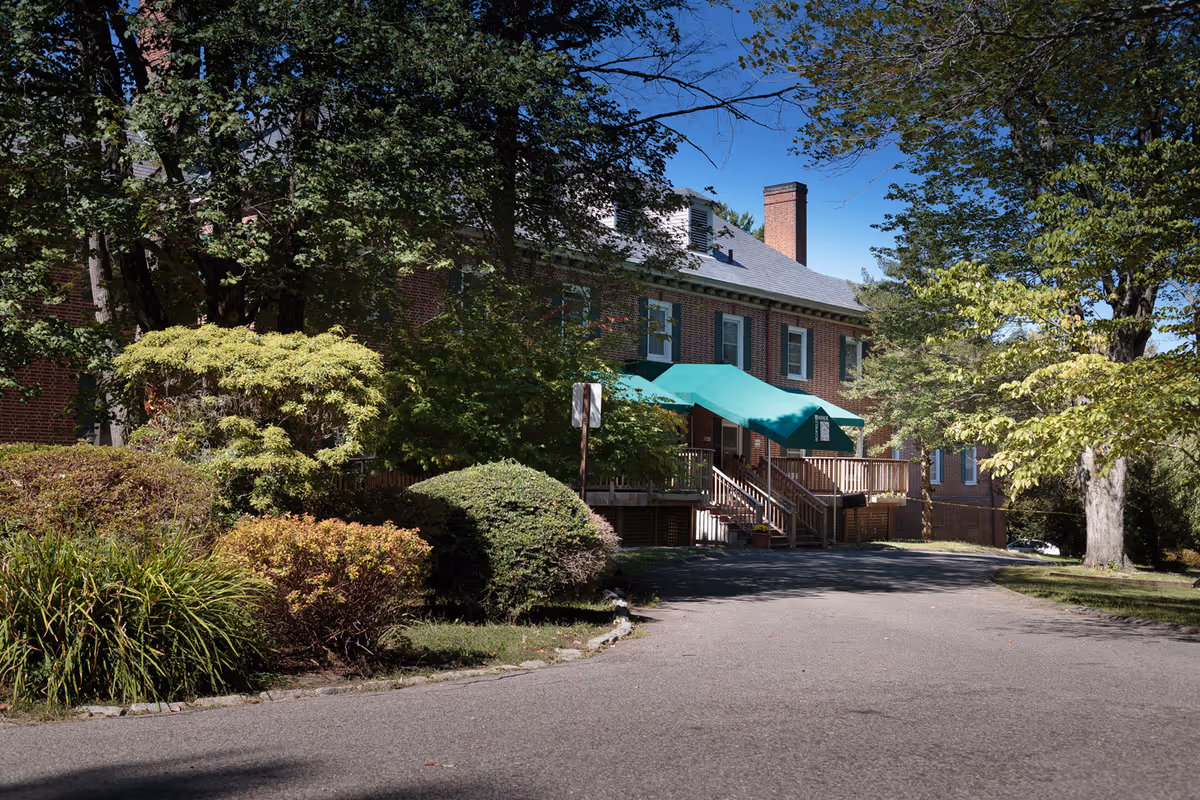 Brick two-story building with a green awning over wooden steps surrounded by trees and shrubs.