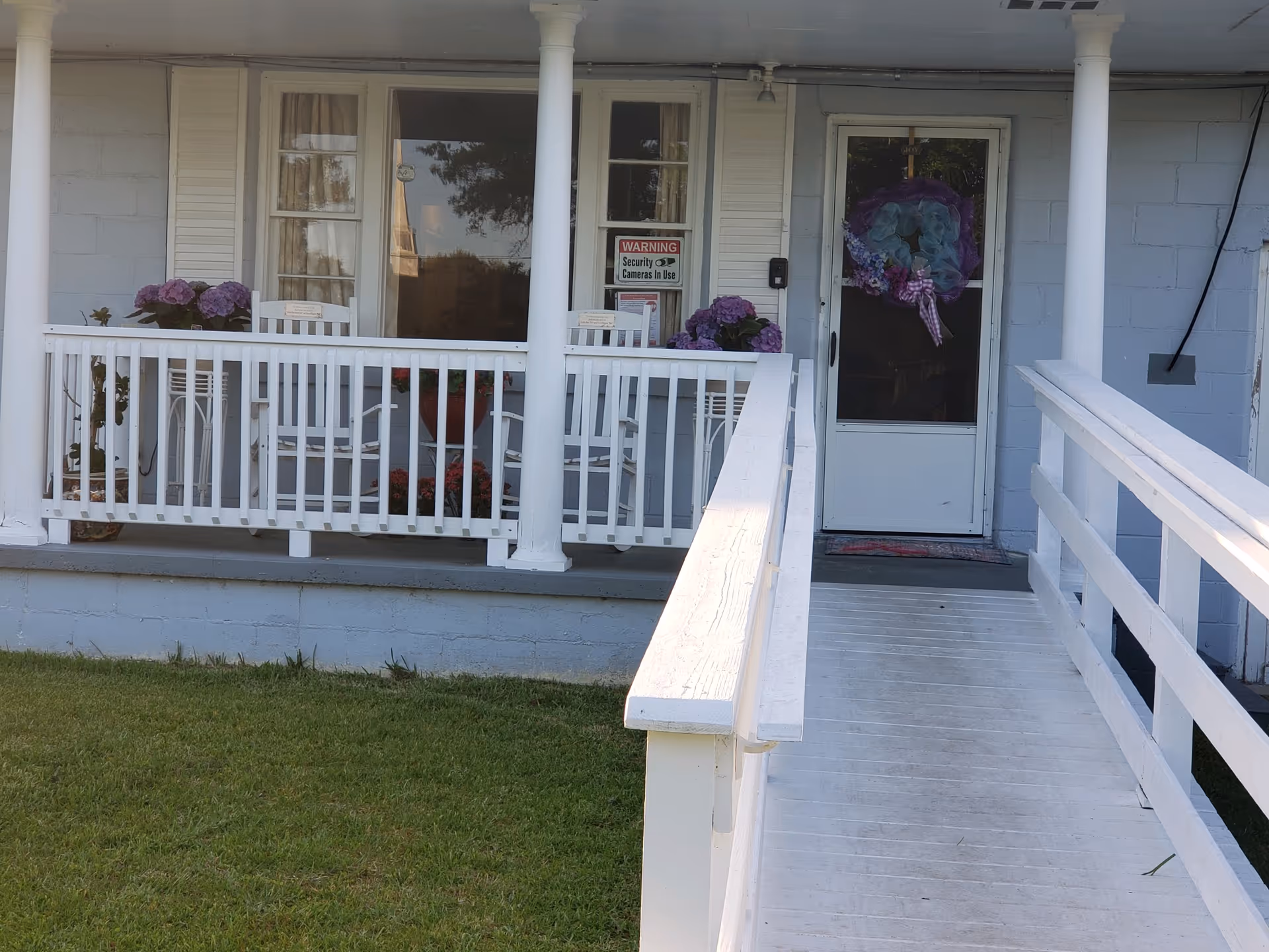 Front porch of The Rhema House Assisted Living Facility, Inc. featuring a white wooden ramp with railings leading to a white door decorated with a colorful wreath. The porch has white railings, two white rocking chairs, potted flowers, and two windows with curtains. A security warning sign is visible on the window next to the door.