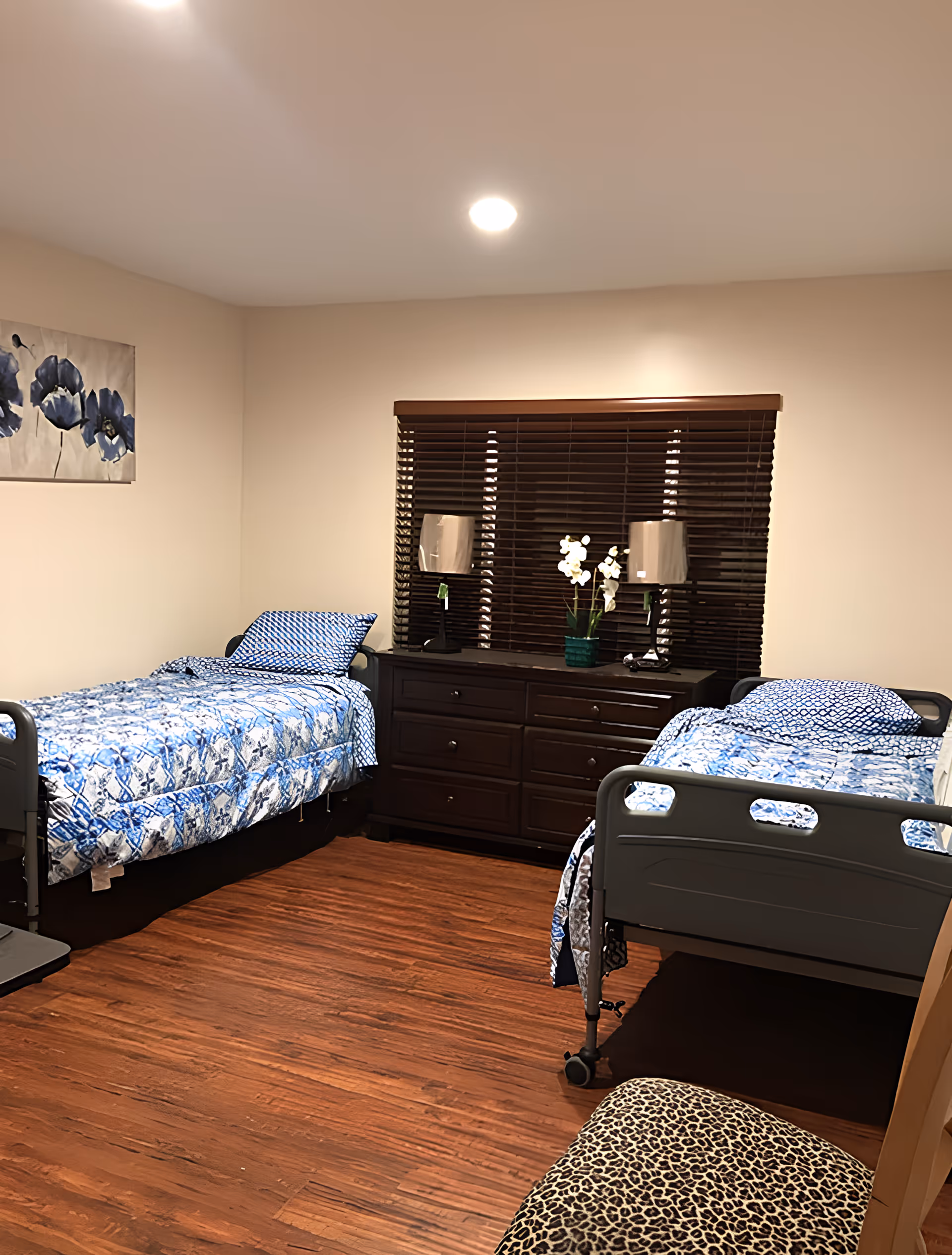 Shared bedroom with two adjustable twin beds dressed in blue patterned bedding, a dark wood dresser under a window with blinds, and hardwood floors.