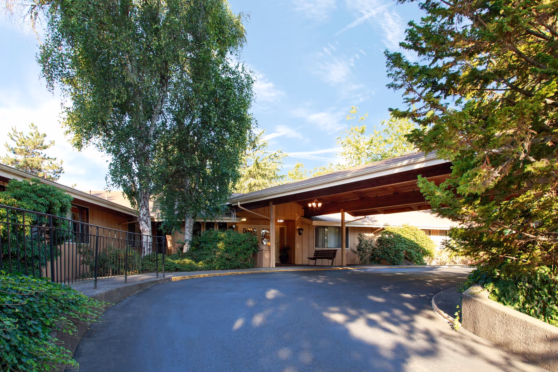 Covered porte-cochere and circular driveway leading to the entrance of a single-story building surrounded by trees and shrubs.