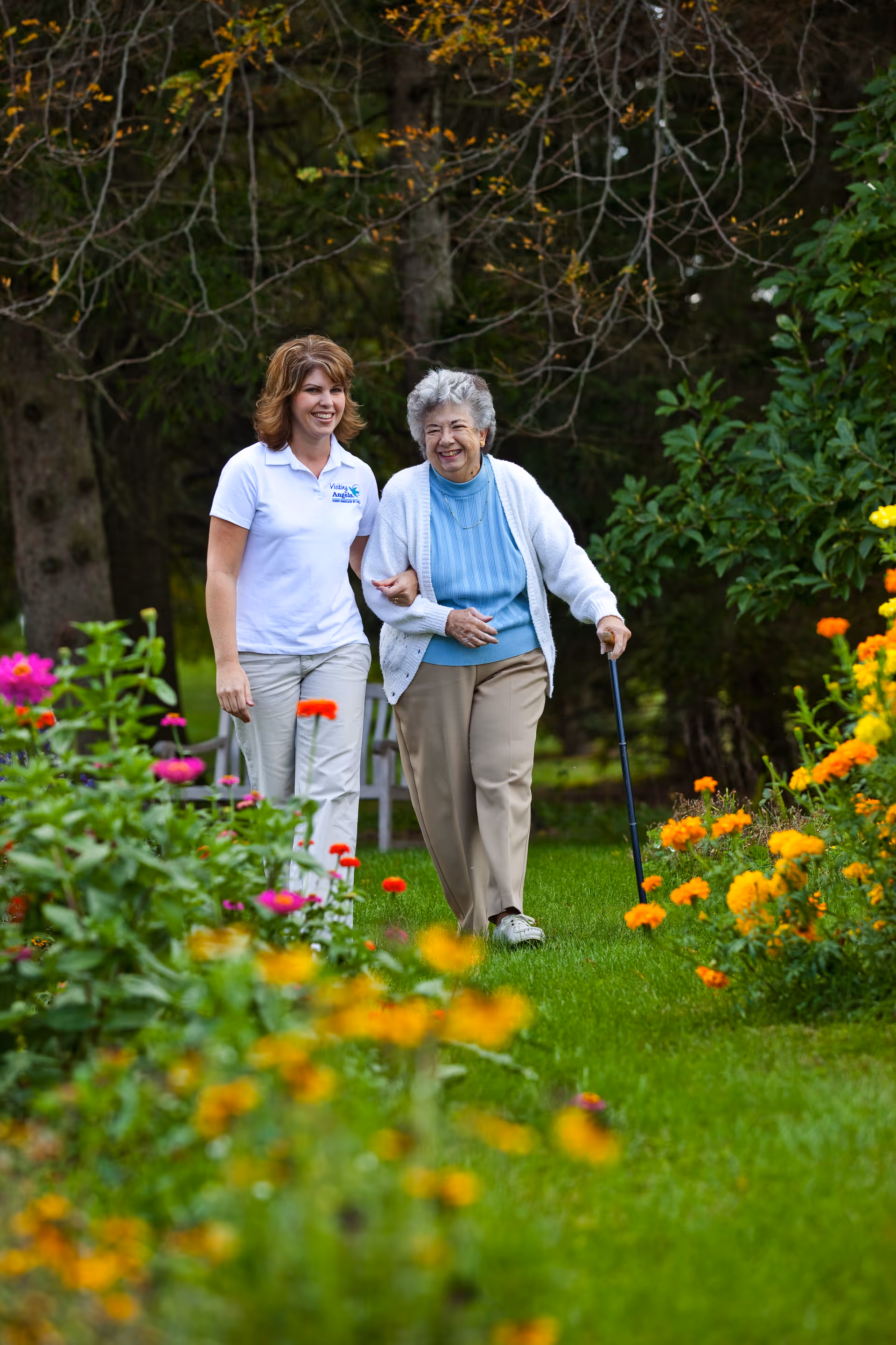 Caregiver assists an elderly woman walking in a sunny garden with colorful flowers.