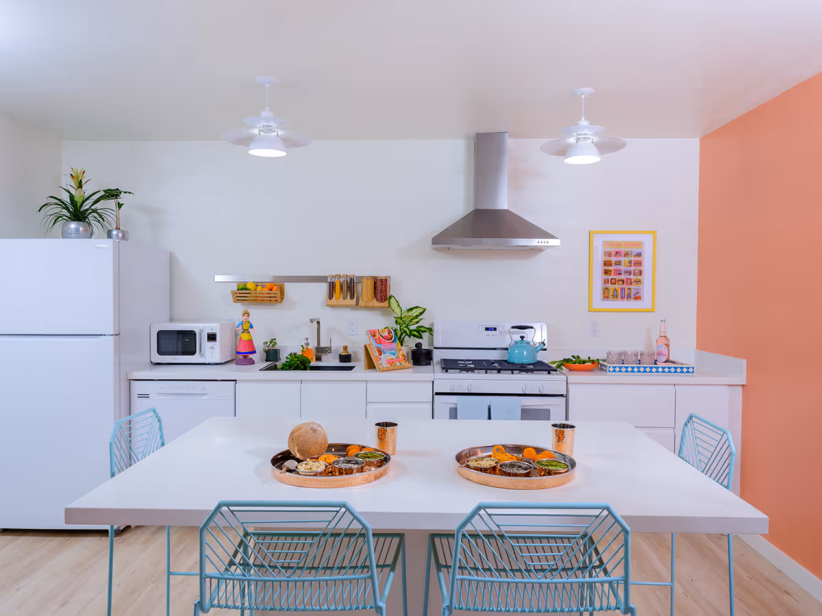 Modern kitchen with white cabinets and appliances including a refrigerator, microwave, and stove with a stainless steel range hood. A white dining table with four blue wireframe chairs is set with two trays of food and copper cups. The kitchen has a white wall with a small framed picture and a peach-colored accent wall on the right. There are plants and kitchen accessories on the countertop.