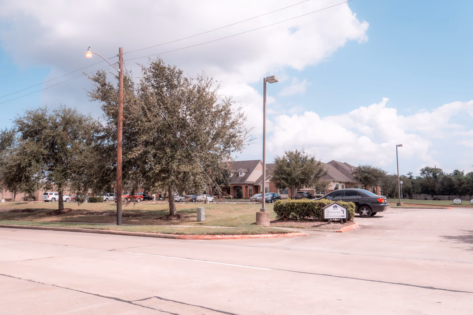 Exterior view of a one-story senior living building with cars parked, trees, lamp posts, and a small sign near the driveway.
