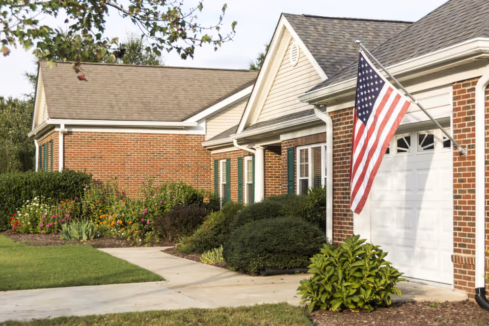 Exterior view of a brick building with a white garage door and an American flag mounted on the wall. There are green bushes and colorful flowers along the sidewalk leading to the entrance.