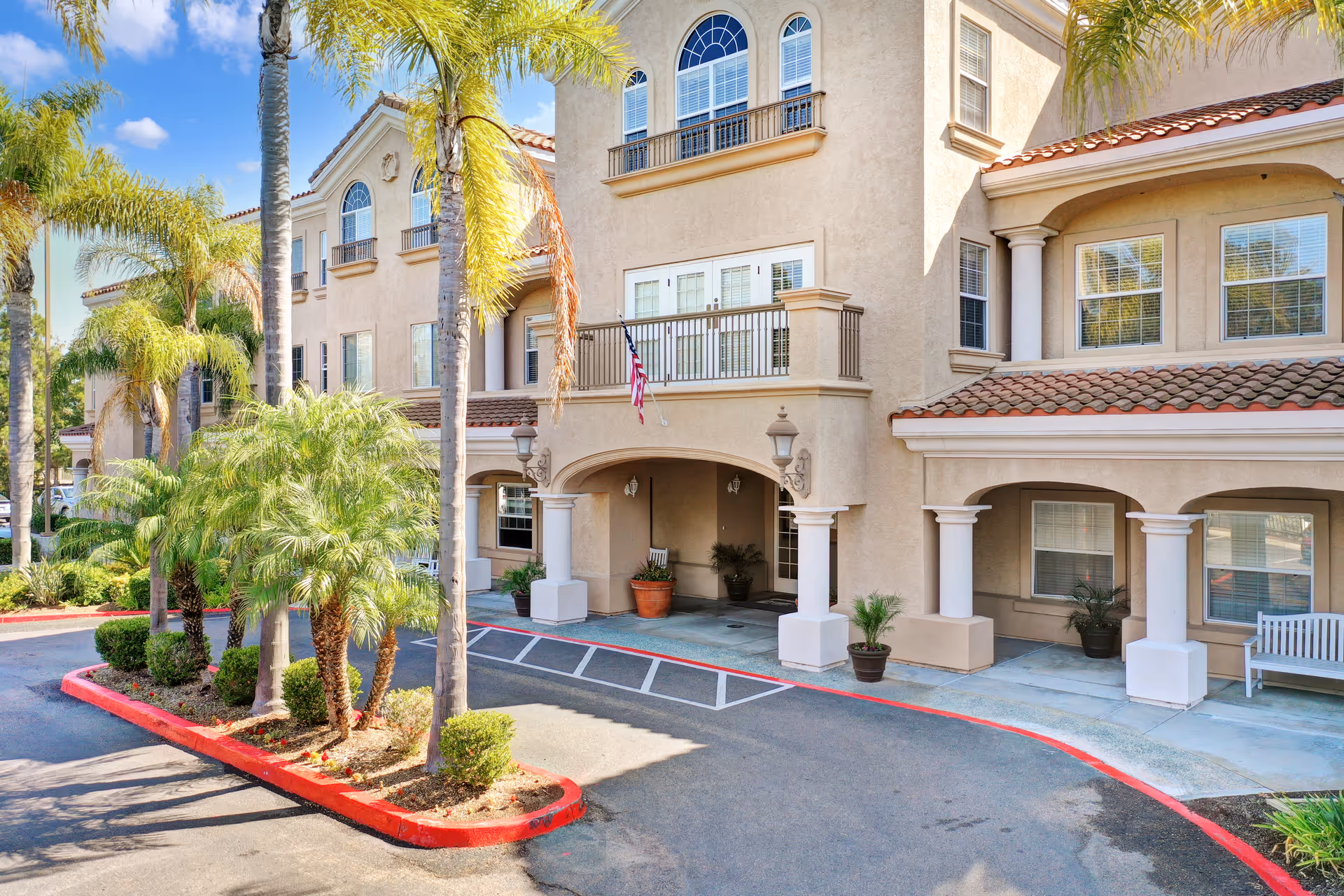 Two-story tan senior living building with arches, columns, balconies, and palm trees by a driveway.