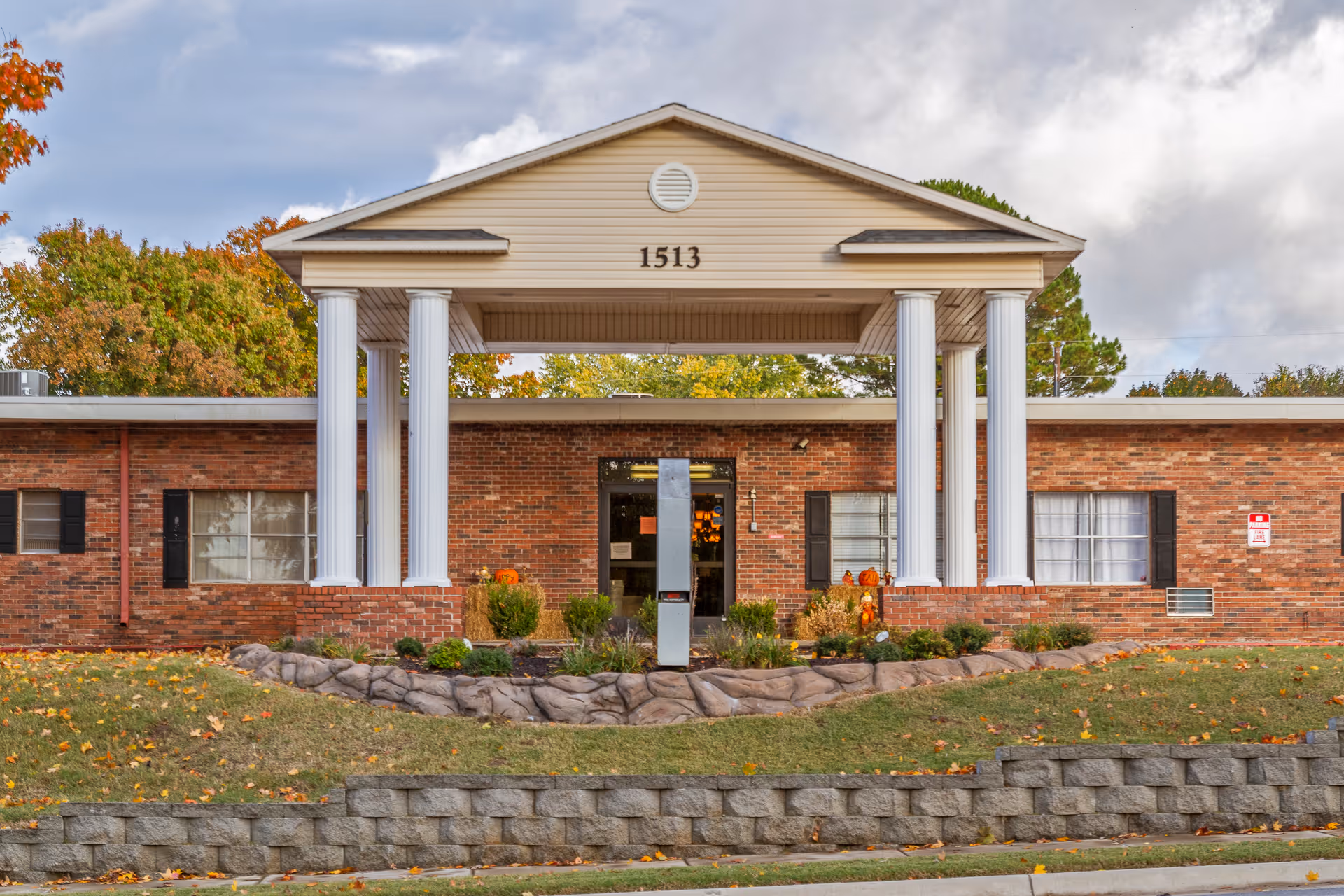 Front exterior view of The Blossoms at Rogers Rehab & Nursing Center, a single-story brick building with white columns supporting a triangular pediment displaying the number 1513. The entrance is surrounded by landscaping with shrubs and autumn decorations, and trees with fall foliage are visible in the background.