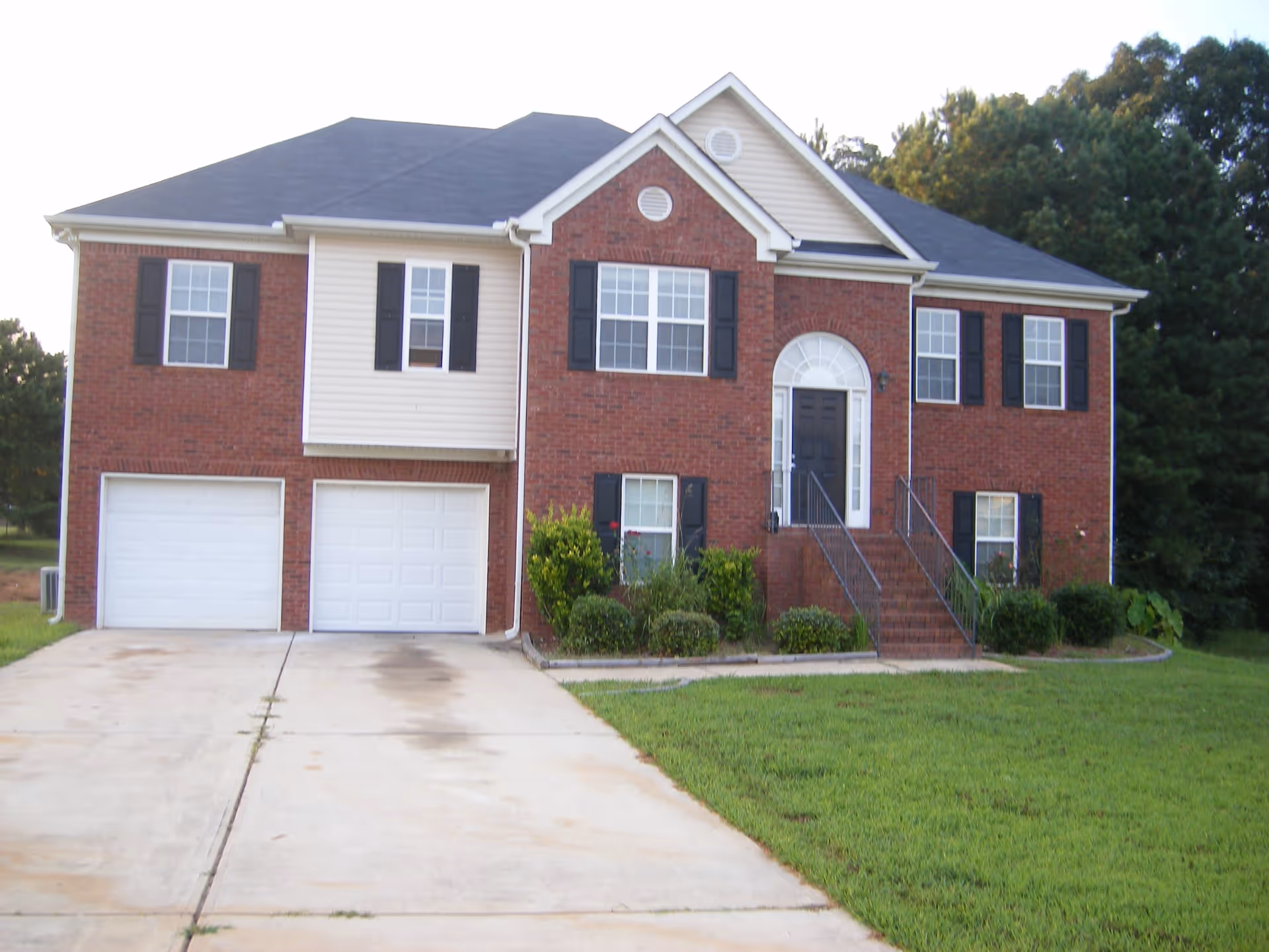 Front exterior view of a two-story brick and siding house with a black front door, multiple windows with black shutters, a double garage, a concrete driveway, and a well-maintained lawn with shrubs and trees in the background.