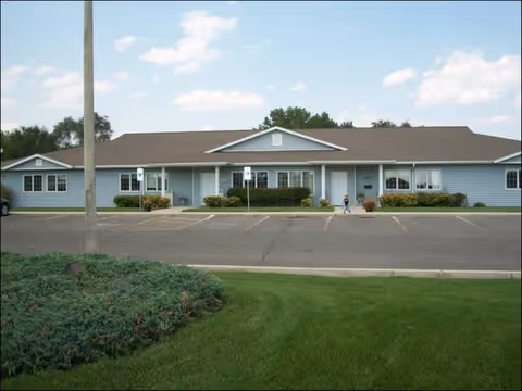 Single-story building with light blue siding and a brown roof, surrounded by a parking lot and green lawn under a partly cloudy sky. A person is walking near the entrance.