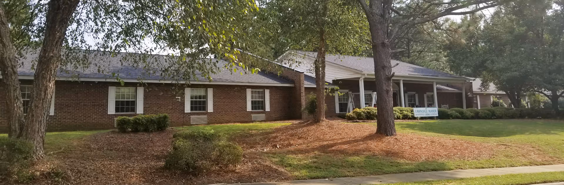Single-story brick assisted living building front with trees, lawn, and a small entrance sign reading 'Lawndale Manor'.