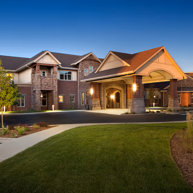 Exterior view of a senior living facility building at dusk with a covered entrance supported by stone pillars, well-maintained green lawn, and landscaped plants.