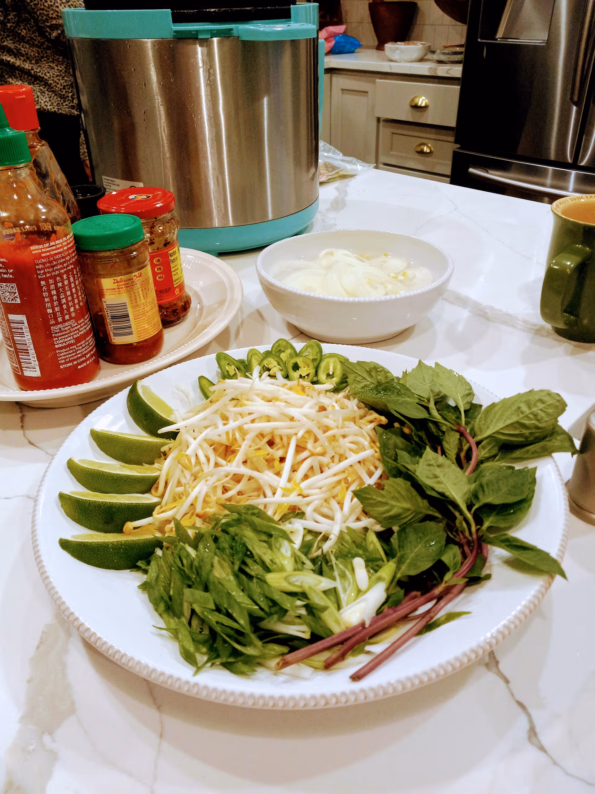 A plate of fresh herbs, bean sprouts, lime wedges, and jalapeños sits on a kitchen counter.