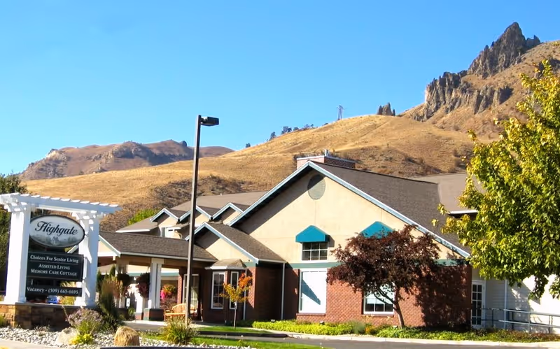 Exterior view of Highgate Senior Living facility with a mountainous backdrop. The building has a beige and brick facade with teal awnings over the windows. There is a white sign in front displaying the facility name and contact information, surrounded by landscaping including trees and shrubs.