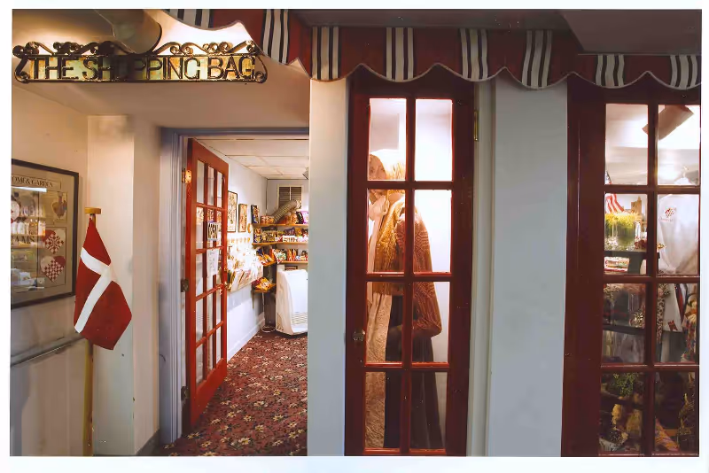 Interior view of a small shop area named 'The Shopping Bag' with red-framed glass doors and a striped awning above. Inside, shelves are stocked with various items. A Danish flag is displayed on a stand to the left, and a mannequin dressed in traditional clothing is visible through one of the glass doors.