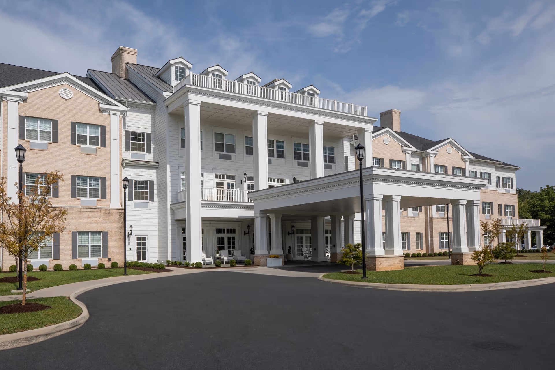 Exterior view of a large, three-story senior living facility building with a covered entrance supported by white columns. The building features a combination of light brick and white siding, multiple windows with shutters, and a paved driveway leading to the entrance. There are small trees and landscaped areas around the building under a partly cloudy sky.