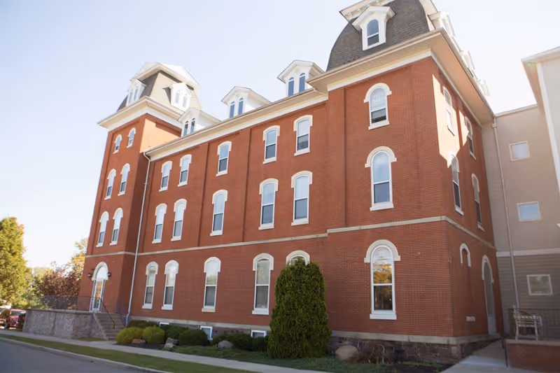 Red-brick, multi-story building with arched windows, dormer roofs, and a small landscaped front.