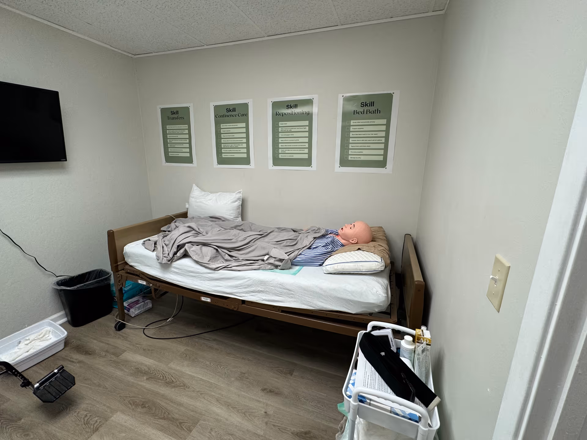 Hospital-style bed in a small training room with a mannequin and wall posters.