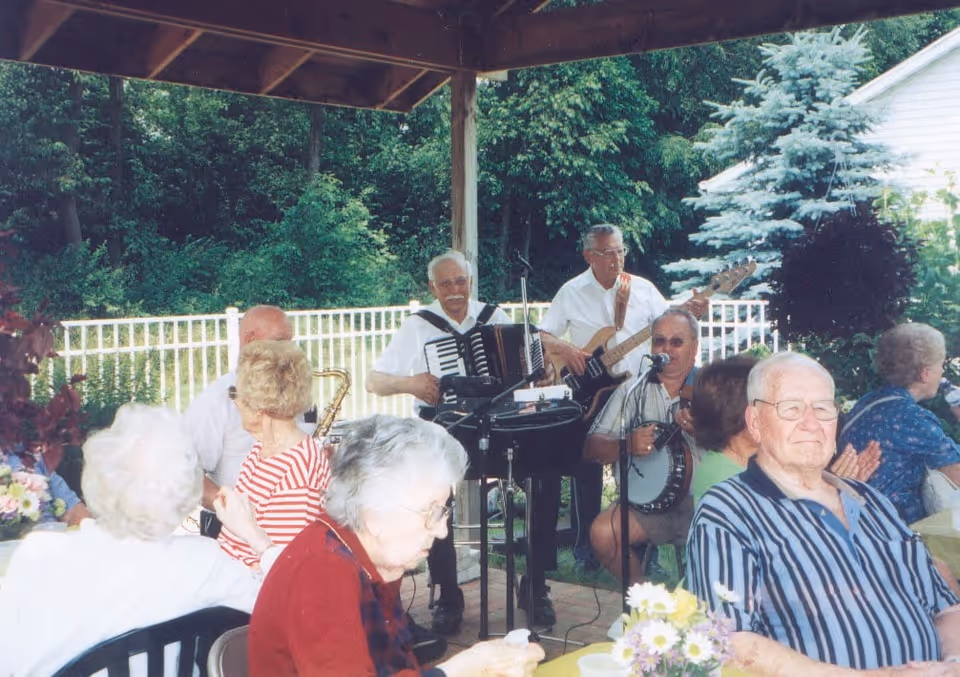 A group of elderly people seated outdoors under a covered patio, enjoying a live band performance. The band consists of three older men playing an accordion, a bass guitar, and a banjo. The setting is surrounded by greenery and a white fence.