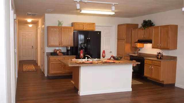 Open-concept kitchen with island, wooden cabinets, and black appliances in a senior living facility.