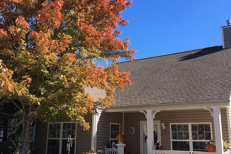 Exterior view of a single-story building with beige siding and a gray shingled roof under a clear blue sky. A large tree with autumn-colored leaves stands to the left, partially shading the building's front porch area, which has white columns and decorative items near the entrance.