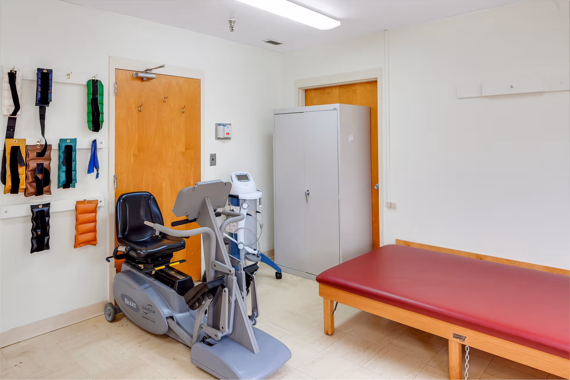 Exercise equipment, a therapy table, cabinets, and wall-mounted accessories in a clinical room.