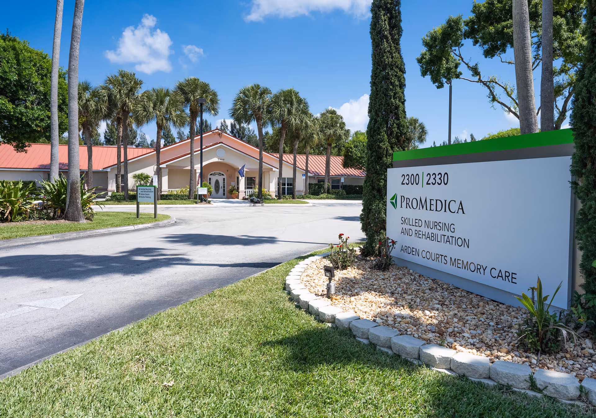 Front entrance of the Arden Courts ProMedica Memory Care building with a driveway, palm trees, and a large sign displaying the facility name and address.