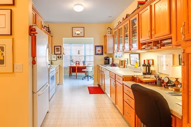 A bright kitchen area with wooden cabinets on the right side and white appliances on the left. The kitchen leads to a sunlit room with a desk and office chair near a large window. The countertop on the right has various items including wine bottles, a lamp, and office supplies. The floor has a light-colored patterned linoleum.