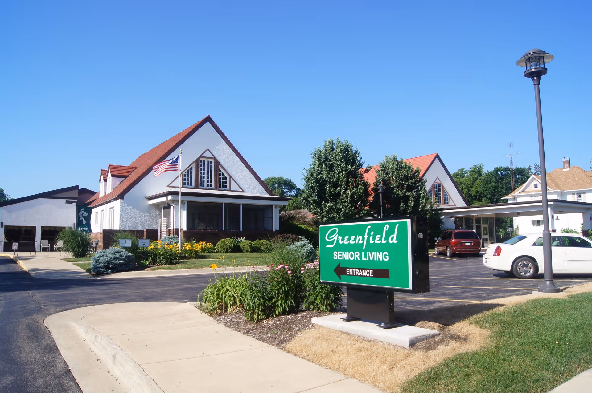 Front exterior of Greenfield Senior Living with a green entrance sign, parking lot, and a white building with red roof under a clear blue sky.