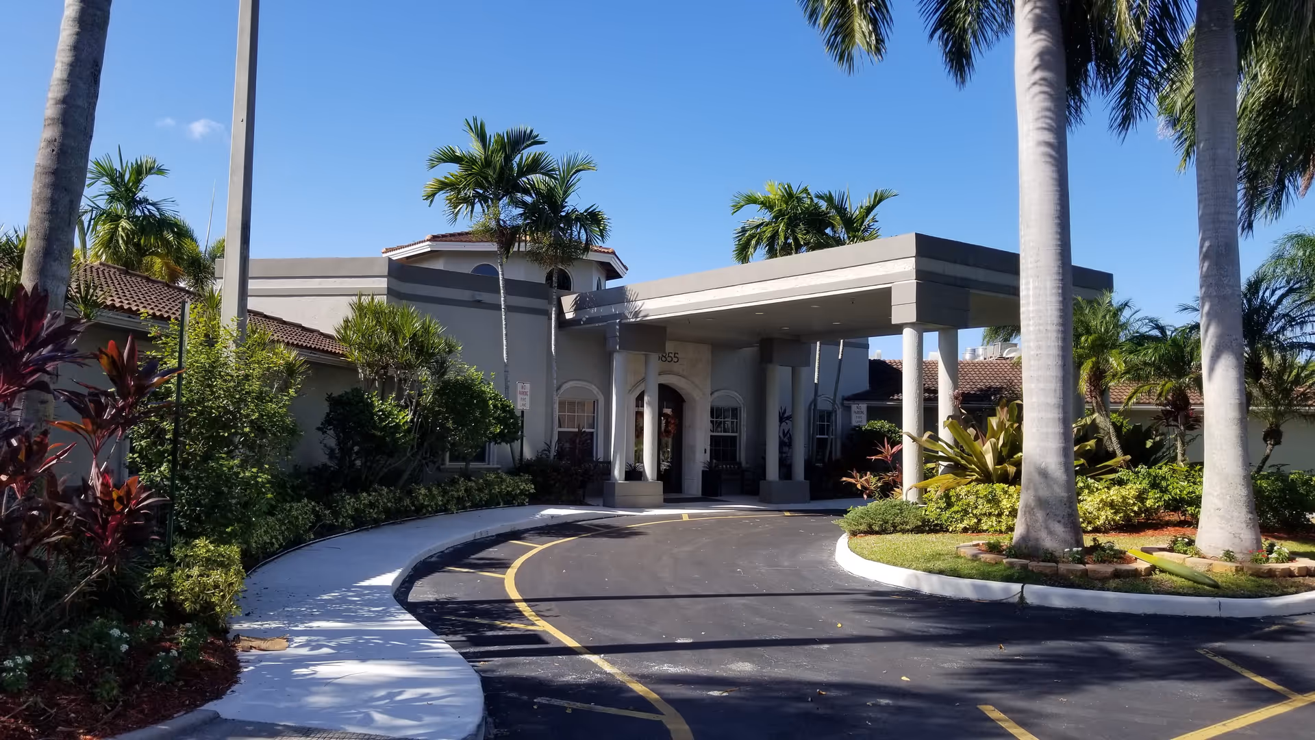 Exterior view of HarborChase of Tamarac entrance with palm trees and a curved driveway.