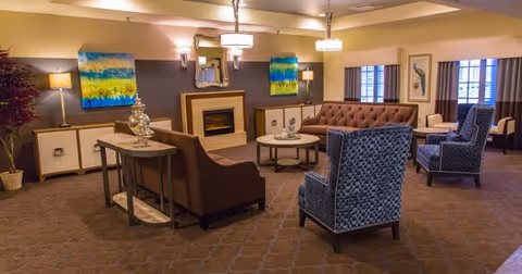 A cozy communal lounge with sofas, patterned armchairs, a central coffee table, fireplace, and colorful wall art.