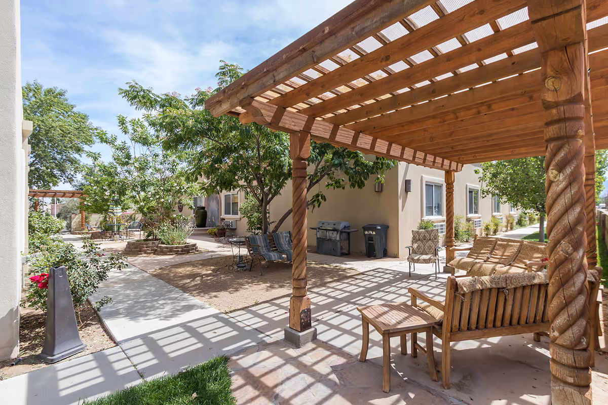 Outdoor patio area at The Heritage Assisted Living & Adult Day Care, LLC featuring a wooden pergola with seating including cushioned chairs and a bench. There are trees and plants around the paved walkway, and a barbecue grill is visible near the building.
