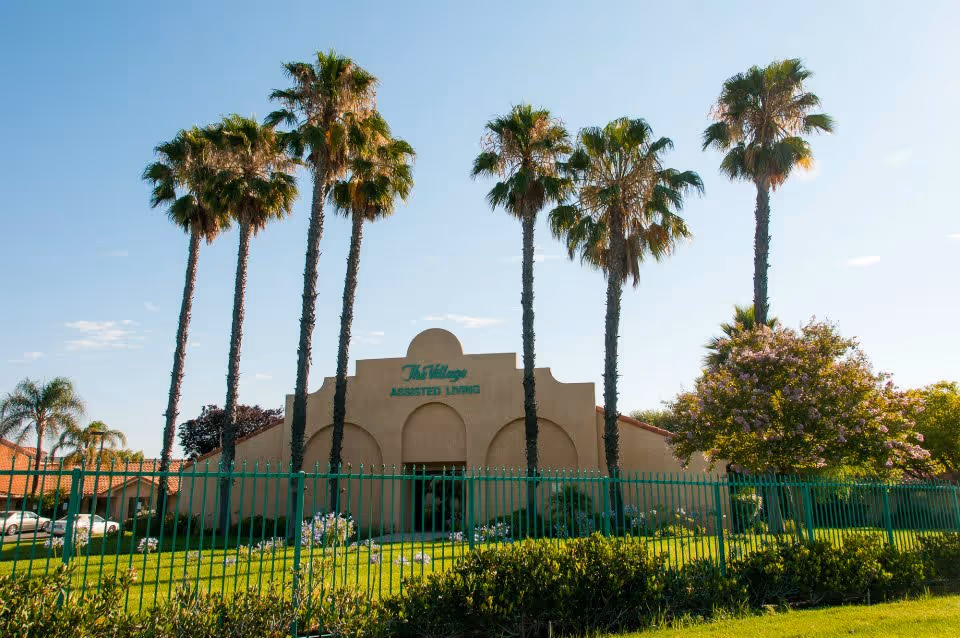 Front exterior of The Village assisted living building with tall palm trees and a green fence.