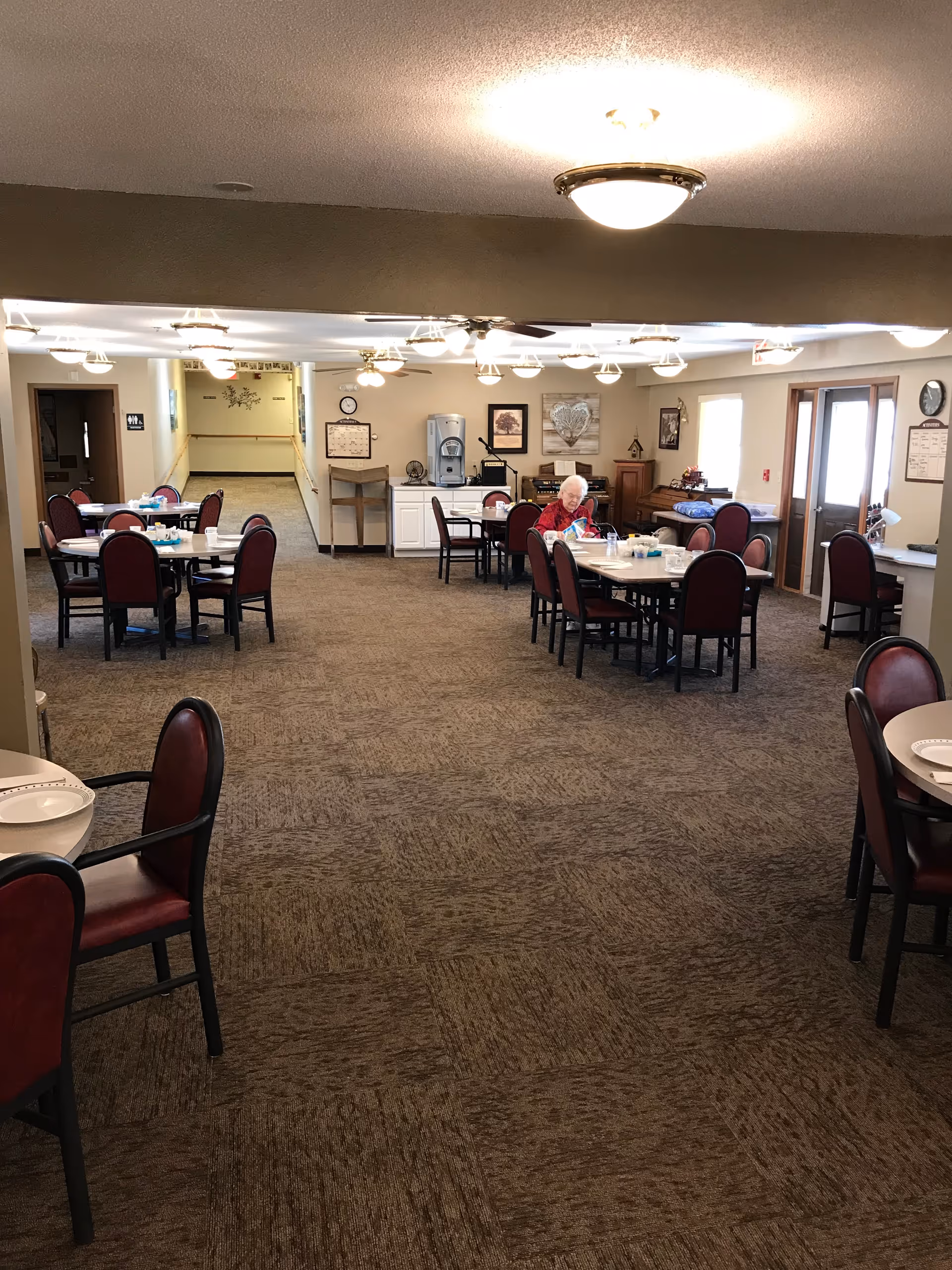 A spacious dining room in an assisted living facility with several round tables and chairs arranged neatly. An elderly woman is seated at one of the tables near the center, reading or looking at something on the table. The room is well-lit with ceiling lights and has a carpeted floor. There is a water dispenser and some framed artwork on the far wall, along with a piano and other furniture.