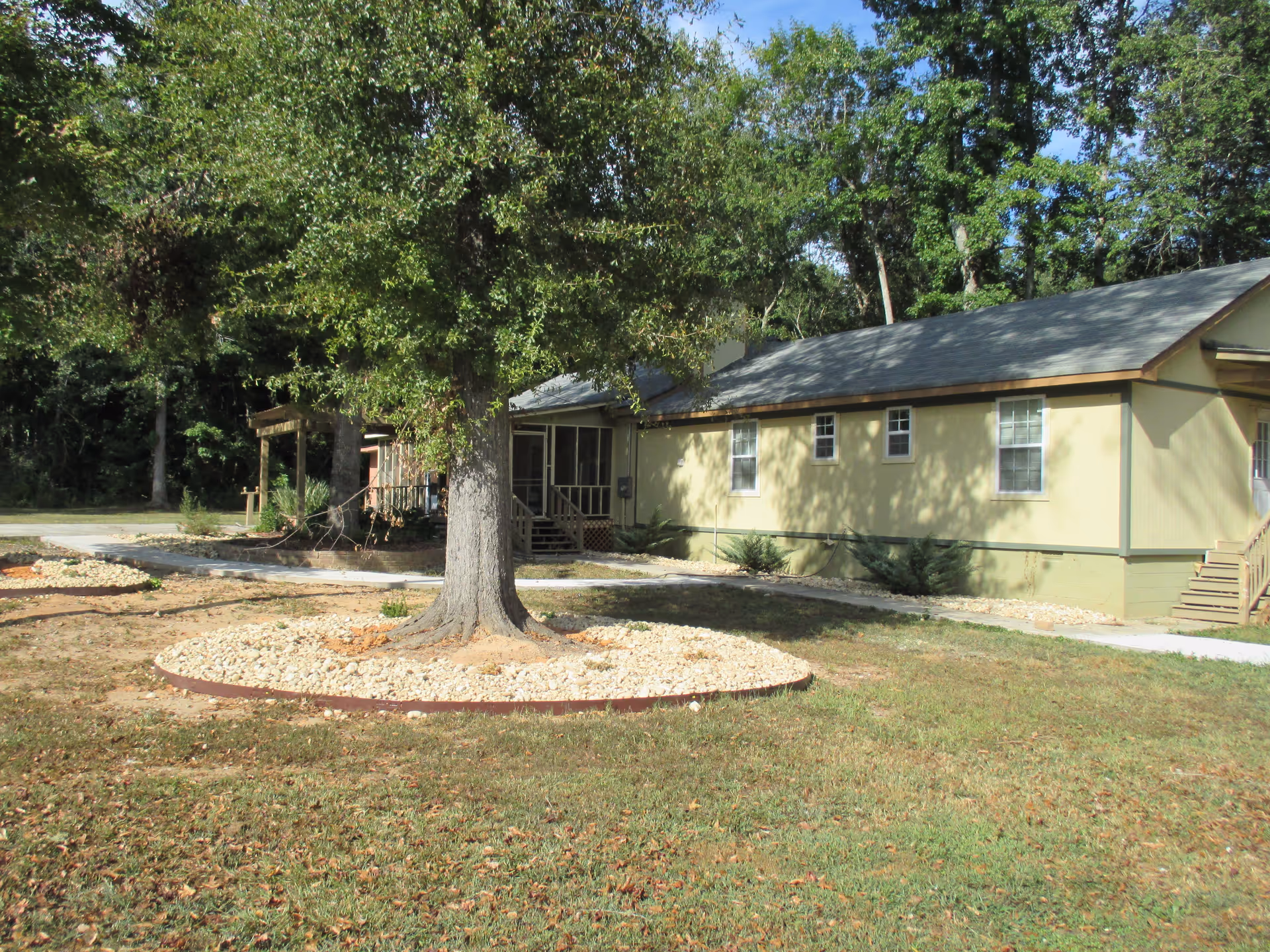 A single-story beige building with a screened porch, surrounded by a lawn, a large tree and circular rock landscaping.
