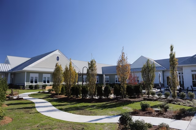 Single-story senior living facility exterior with curved walkway, landscaping, and blue sky.