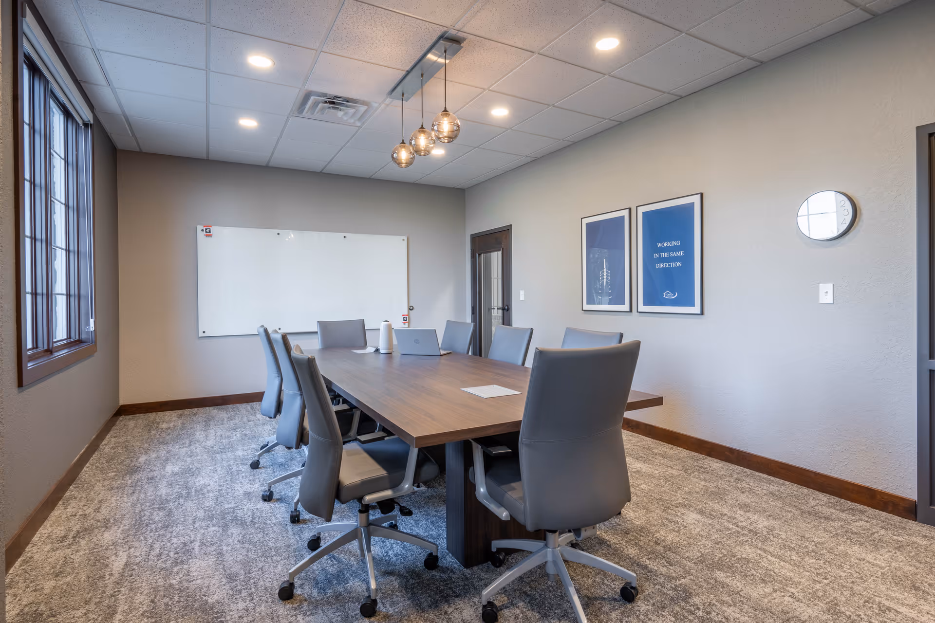 Conference room with long wooden table and grey chairs, whiteboard, windows, and framed art.