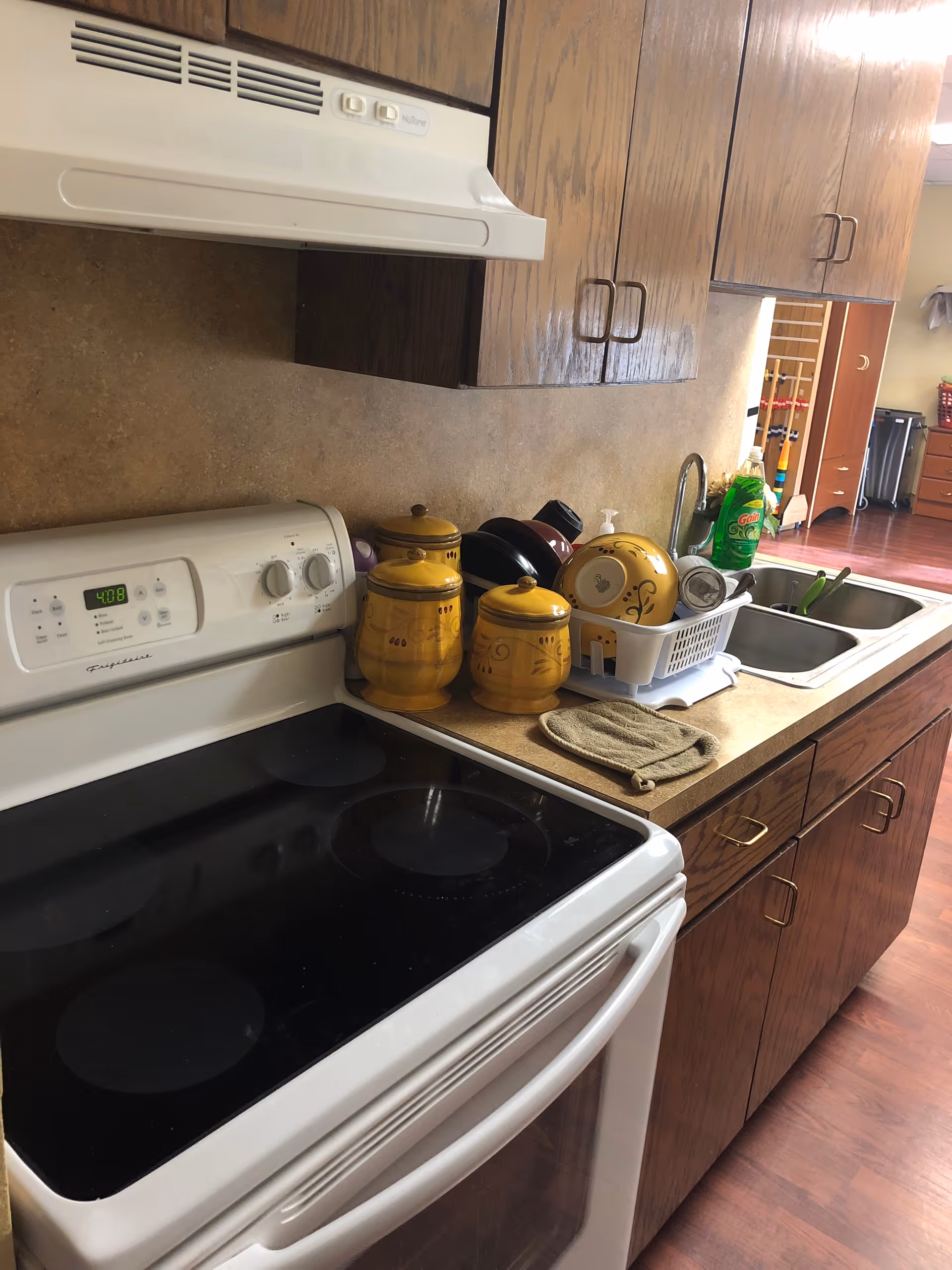 A kitchen area with a white electric stove and oven, wooden cabinets above and below the countertop, a double sink with a dish rack holding dishes and utensils, and three yellow ceramic canisters on the counter. There is a bottle of green dish soap near the sink and a beige oven mitt on the counter.