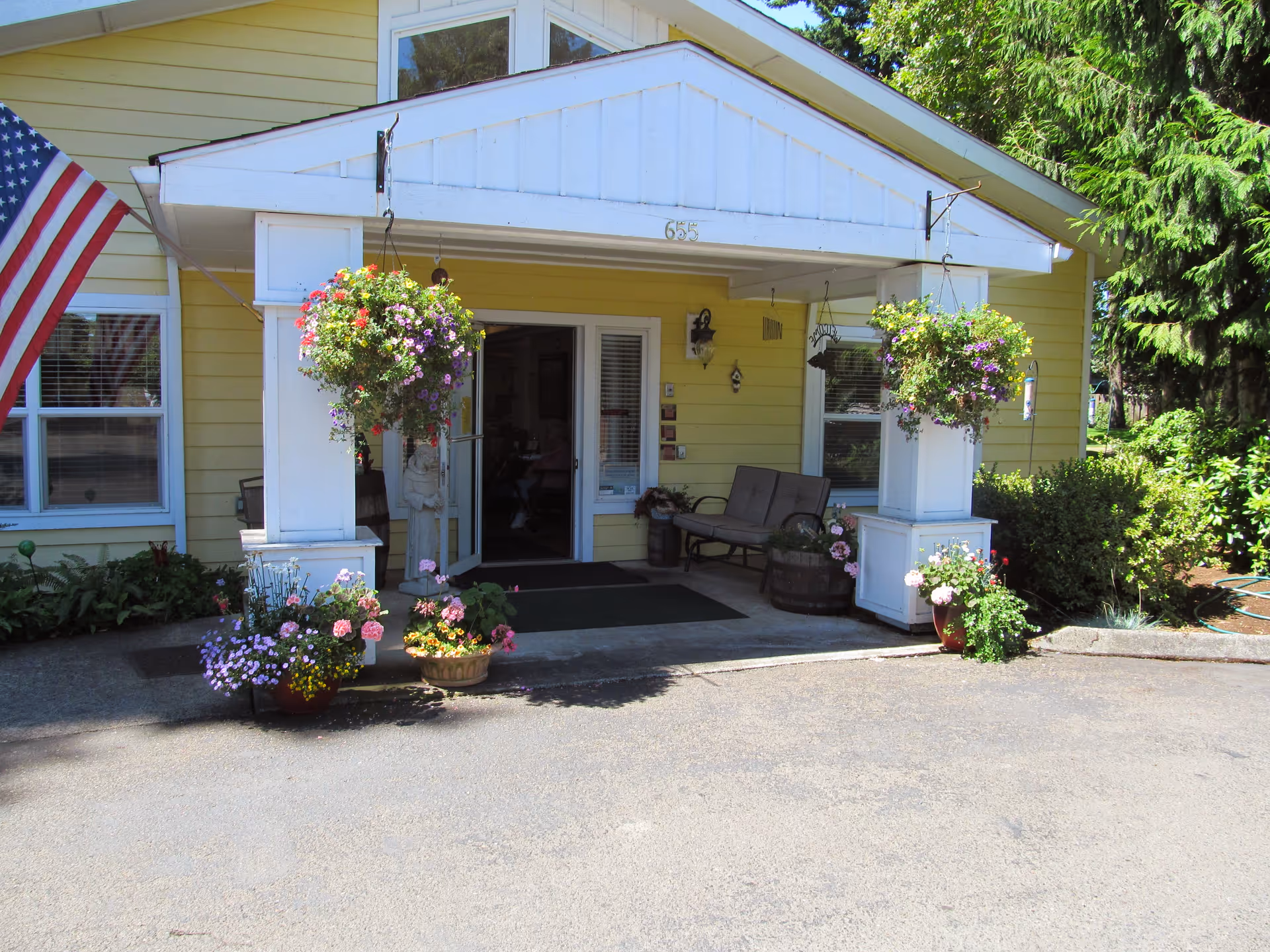 Small yellow facility entrance with hanging flower baskets, porch seating, and an American flag.