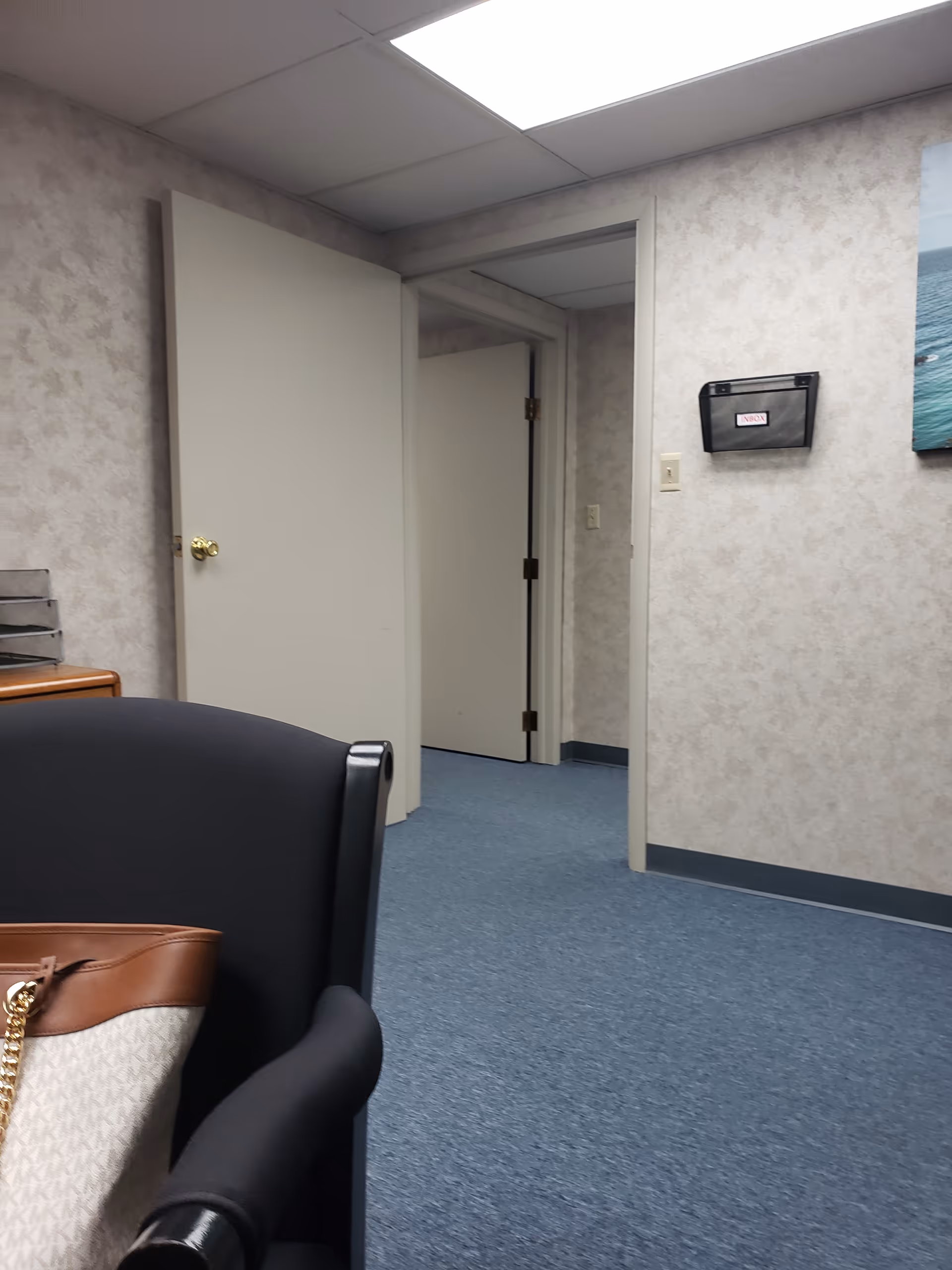 Indoor facility hallway with blue carpet, a foreground chair, and open doors.
