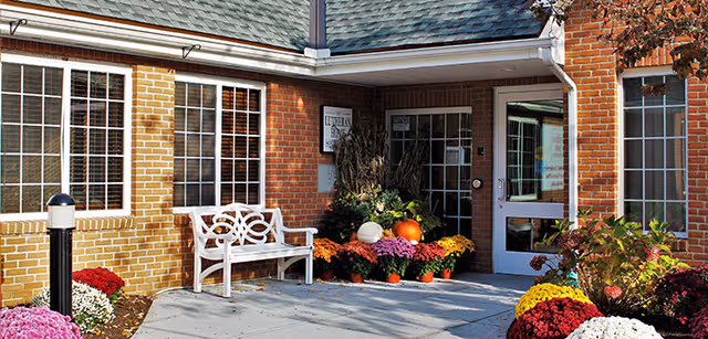 Brick entrance of a senior living facility with a white bench, potted chrysanthemums and pumpkins by the doorway.